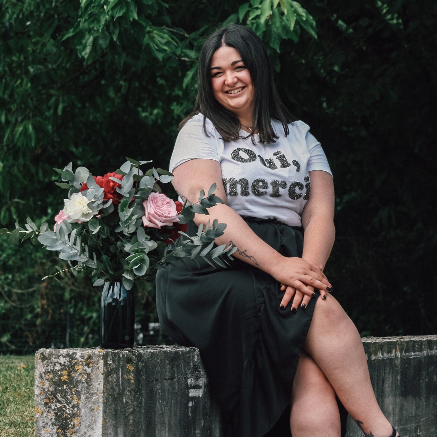 Jeune fille brune qui est assise à côté d'un vase réalisé dans une bouteille de champagne. Le vase est rempli de jolies fleurs. 