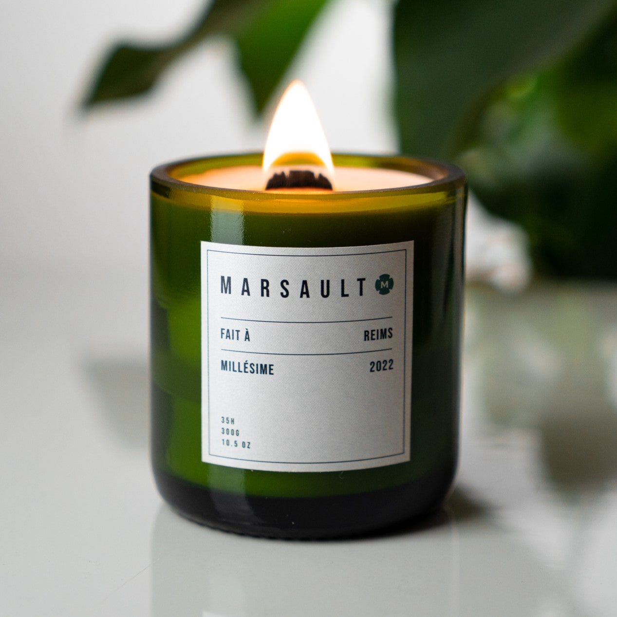 Bougie Marsault verte format bouteille de champagne qui est allumée à l'intérieur devant une plante verte.